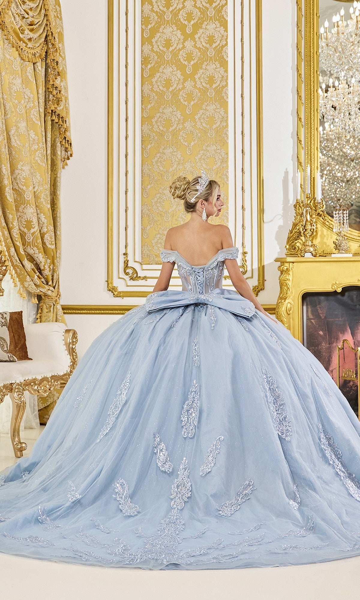 A woman in the Dusty Blue Quinceanera Dress 5089 by Dancing Queen, featuring off-the-shoulder lace applique, stands with her back to the camera in an ornate gold-decorated room.